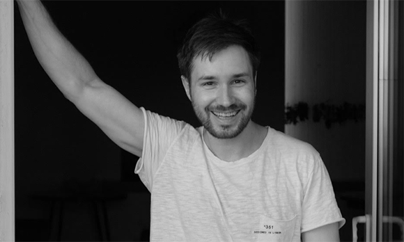 A smiling man in a white t-shirt raises one arm against a dark background.