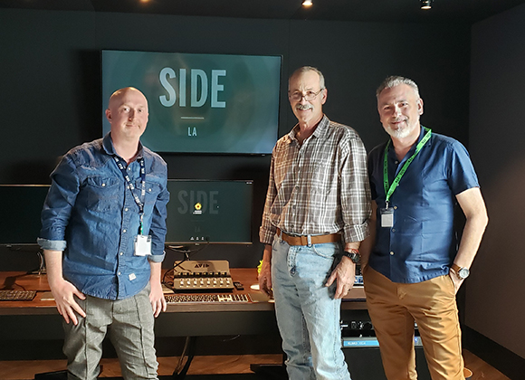 Three men stand in a studio before a large screen displaying the text SIDE LA.