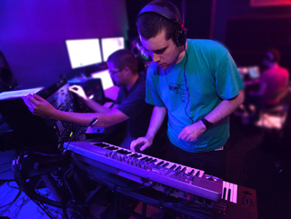 A man in a teal shirt wearing headphones plays a keyboard in a dark studio with blue lighting.