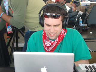 A man in a green shirt and red bandana wears headphones while looking at a laptop.
