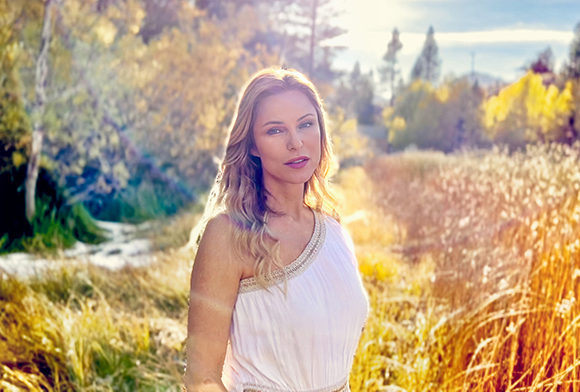 A blonde woman in a white sleeveless dress stands in a golden, sunlit field of tall grass.