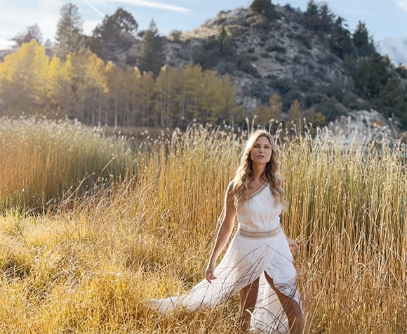 A woman in a white dress stands in a field of tall golden grass with trees and mountains in the background.