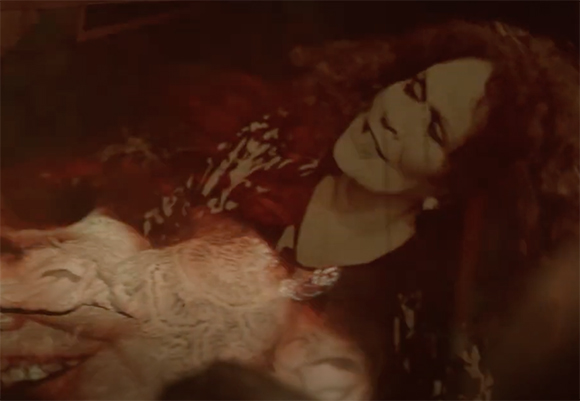 A person with voluminous curly hair lies in a dimly lit, reddish-toned environment.