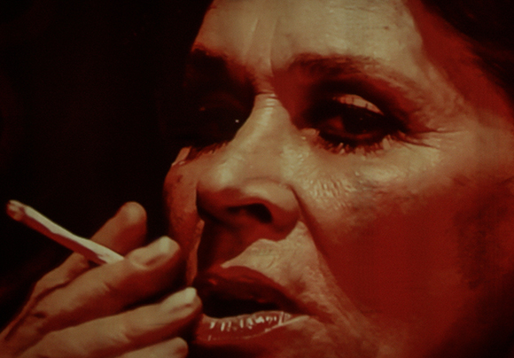 Close-up of a person's face in red lighting while holding a lit cigarette to their lips.