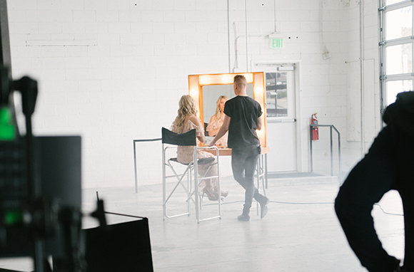 A woman sits at a lighted vanity mirror while a man stands beside her in a bright, white studio space.