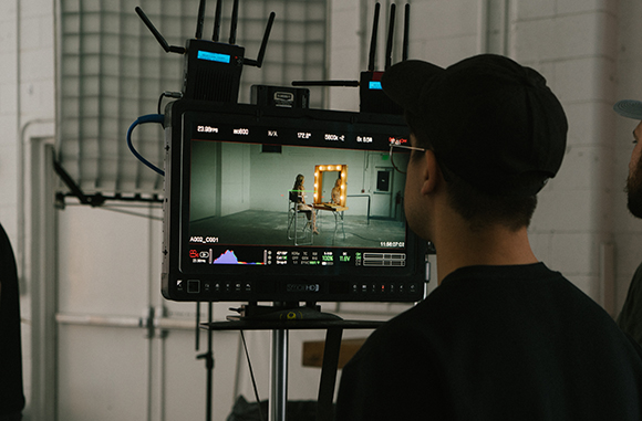 A person in a black cap looks at a professional video monitor displaying a scene of a person sitting at a table.