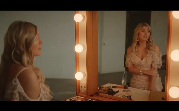 A blonde woman in a white lace dress looks at her reflection in a large vanity mirror framed by glowing bulbs.