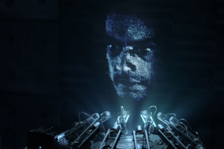 A blue digital projection of a human face above several glowing vacuum tubes in a dark setting.