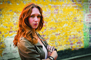 A woman with wavy red hair and a green jacket stands with arms crossed in front of a yellow brick wall.