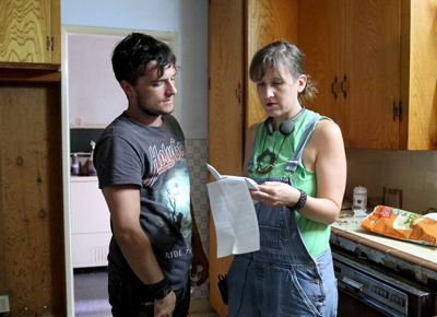 A man in a grey t-shirt and a woman in denim overalls look at a white paper in a kitchen.