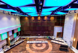 A wide view of a modern performance hall with blue glowing ceiling panels and a wooden stage area.