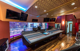A large audio mixing console in a recording studio with wood floors and a view into the live tracking room.