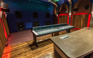 A large audio mixing console sits in a dark studio with blue walls and wood paneling.