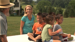 A blonde woman in a teal shirt smiles while sitting with a group of children on a white surface outdoors.