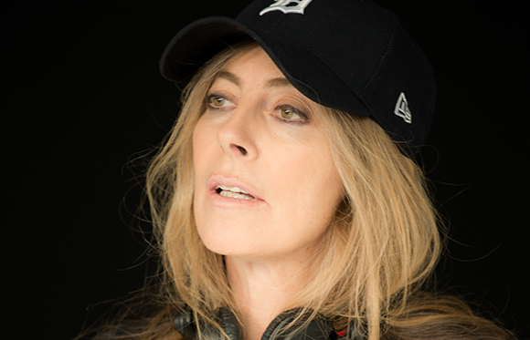 A woman with long blonde hair wearing a black baseball cap looking up against a black background.