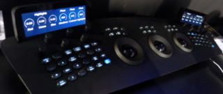 A black control surface with multiple illuminated buttons, knobs, trackballs, and a small screen displaying color grading par