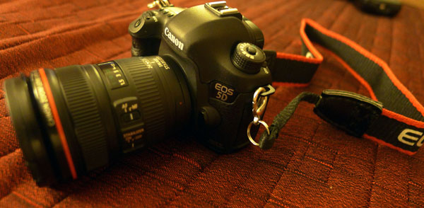 A black Canon EOS 5D camera with a lens and a black and red strap is placed on a textured red surface.