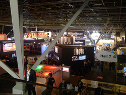 Interior of a large exhibition hall with booths, signage, and people on escalators.