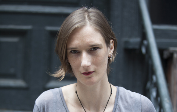 A woman with short brown hair and freckles looks directly at the camera against a dark background.
