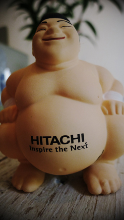 A light-skinned sumo wrestler stress ball with a smiling face and the Hitachi logo on its belly.