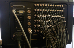 Rear panel of electronic equipment with many BNC connectors, a DB25 connector, and AC power input, with cables attached.