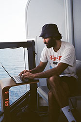 A bearded man in a bucket hat sits by a window and types on a laptop with an ocean view in the background.
