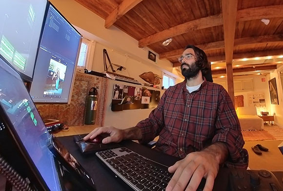 A man with a beard and glasses sits at a desk using a computer and keyboard in a room with wooden beams.