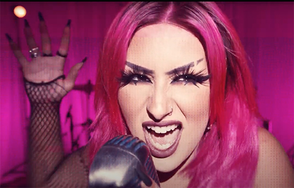 A person with bright pink hair and dramatic eye makeup sings into a microphone.