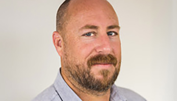 A bald man with a brown beard and blue eyes wearing a light grey collared shirt.