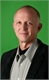 A man with light skin and short hair wearing a black blazer and grey shirt against a green background.