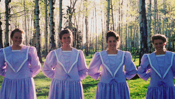 Four women in matching light purple long-sleeved dresses standing in a sunlit forest of thin trees.