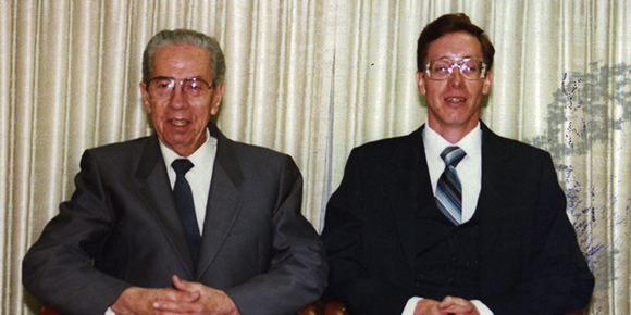 Two men wearing suits and ties sitting in front of light-colored curtains.