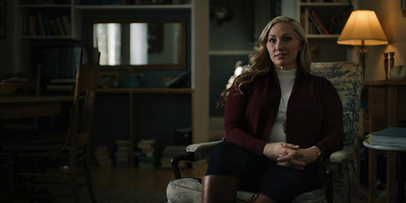 A blonde woman in a maroon cardigan sits in a patterned armchair in a dark living room.