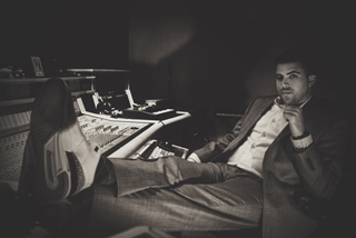 A man in a suit sits reclined in a chair at a large mixing console in a dark studio.