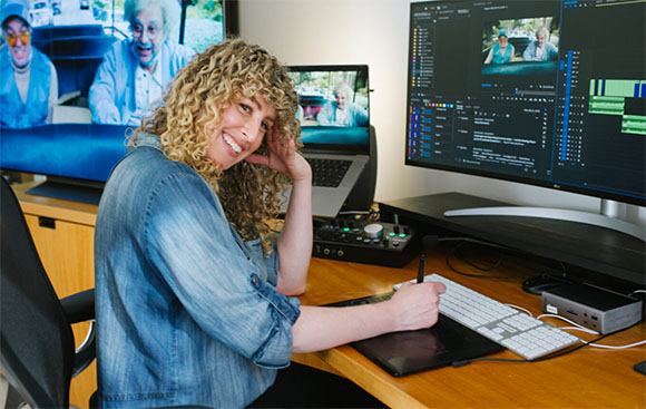 A smiling woman with curly hair uses a digital stylus at a desk with multiple computer monitors.