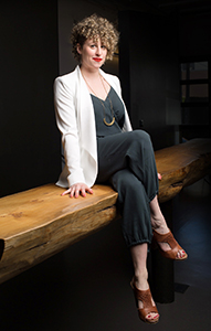 A woman with curly hair wearing a white blazer and dark jumpsuit sits on a rustic wooden bench.