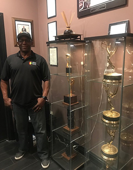 A man in a black polo and cap stands beside two glass display cases filled with gold trophies.