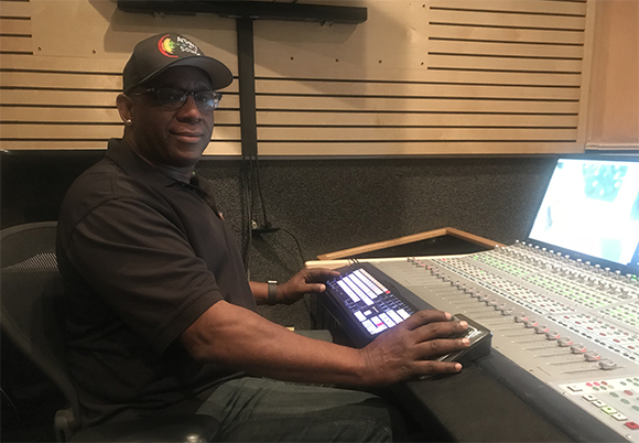 A man in a black polo and cap operates a large audio mixing console in a recording studio.