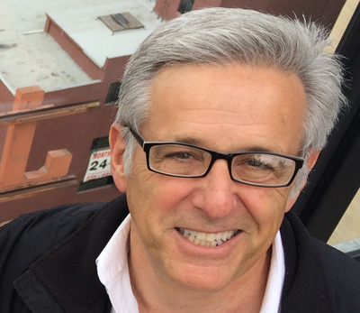 A smiling man with short gray hair and black-rimmed glasses wearing a white shirt and black jacket.