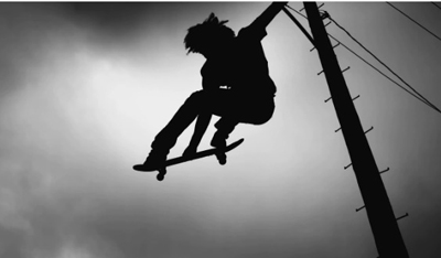 Silhouette of a person mid-air on a skateboard next to a utility pole against a cloudy sky.