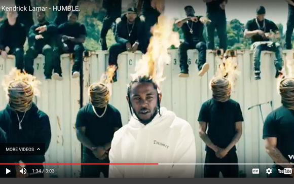 A man in a white hoodie stands in front of several men with cloth-wrapped heads that are on fire.