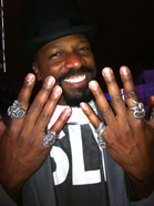 A smiling man wearing a hat and several large, ornate rings on his fingers.