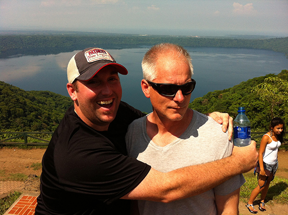Two men embrace outdoors with a large lake and green hills in the background.