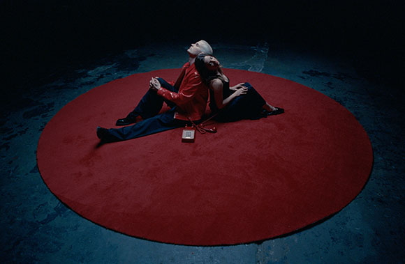 Two people sit back-to-back on a large red circular rug in a dark room.