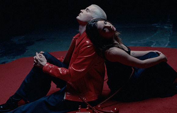 A man in a red jacket and a woman in a black dress sit back-to-back on a red floor against a dark background.