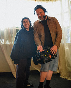 A man wearing headphones and a brown shirt holds a video camera while standing next to a smiling woman.