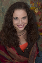 A woman with long, dark curly hair smiles at the camera against a textured background with autumn leaves.