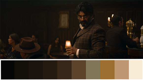 A man with dark hair and a beard wears a plaid suit in a dark, moody room.