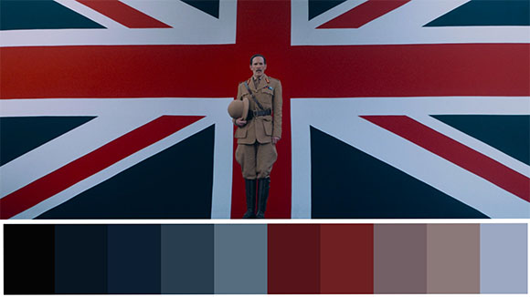 A man in a tan military uniform stands centered before a large British Union Jack flag.