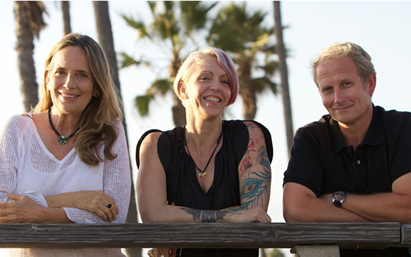 A woman with long blonde hair, a woman with pink hair and tattoos, and a man smiling outdoors.